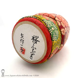 A photo showing the signature of kokeshi artist Kazuko Yahagi. The photo appears on the base of a one-of-a-kind modern kokeshi doll. The doll has a peaceful expression and is made from nicely grained hardwood.