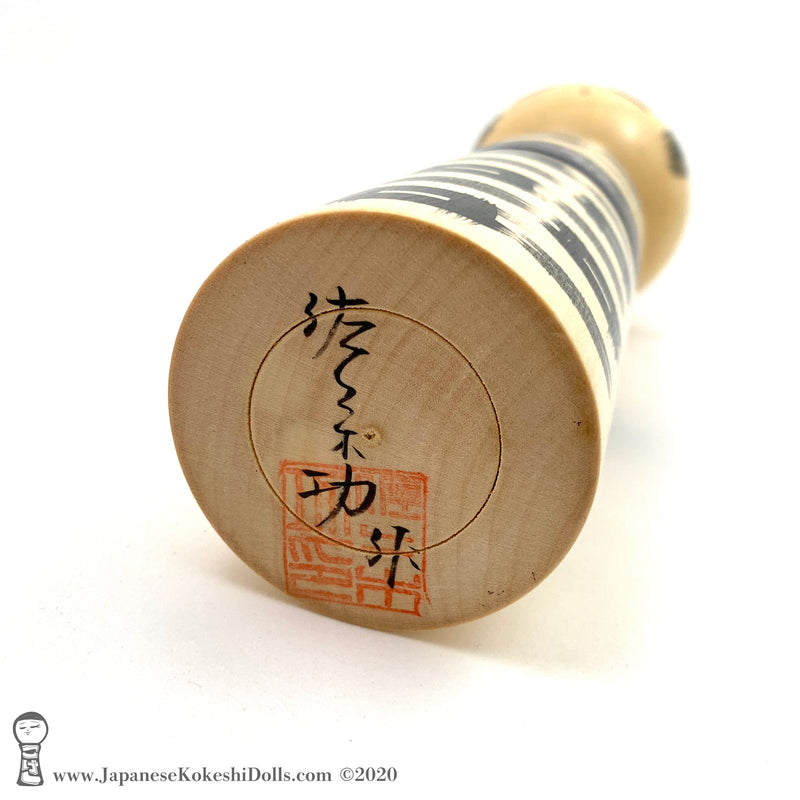 A photo showing the signature of kokeshi artist Isao Sasaki. The photo appears on the base of a one-of-a-kind modern kokeshi doll. The doll has a peaceful expression and is made from nicely grained hardwood.