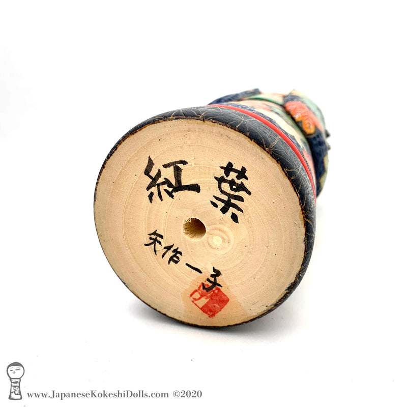 A photo showing the signature of kokeshi artist Kazuko Yahagi. The photo appears on the base of a one-of-a-kind modern kokeshi doll. The doll has a peaceful expression and is made from nicely grained hardwood.